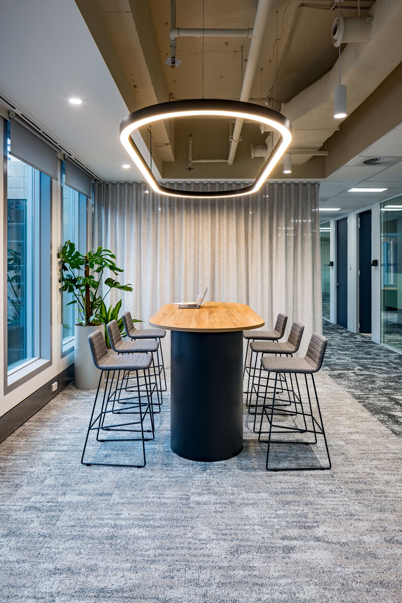 Modern office meeting area with a round wood-top high table, black bar stools, a circular LED pendant light, and sheer curtains.