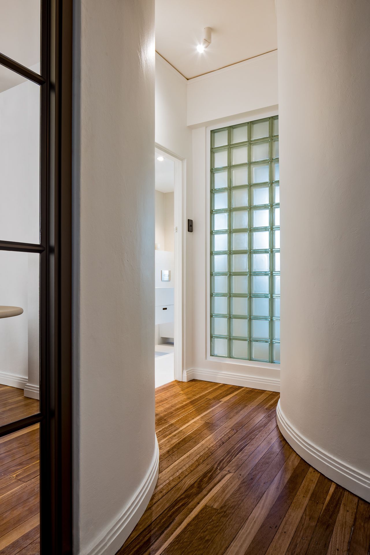 Curved hallway with black-framed glass door and timber floors leading to a bathroom with a glass block window.