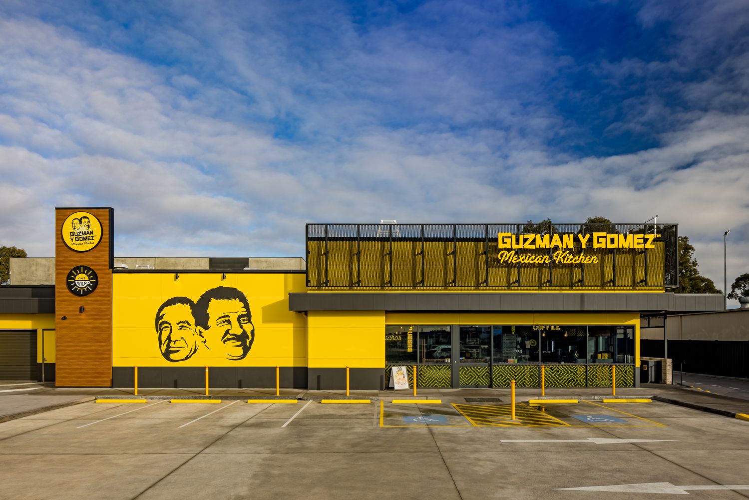Guzman y Gomez Mexican Kitchen drive-through restaurant with yellow facade and the founders' faces on the wall under a cloudy sky.