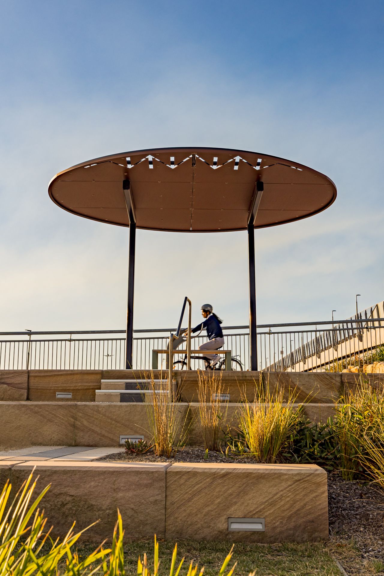 Cyclist rides past a circular rust-colored shade canopy on a landscaped path with stone steps, ornamental grasses and railings.