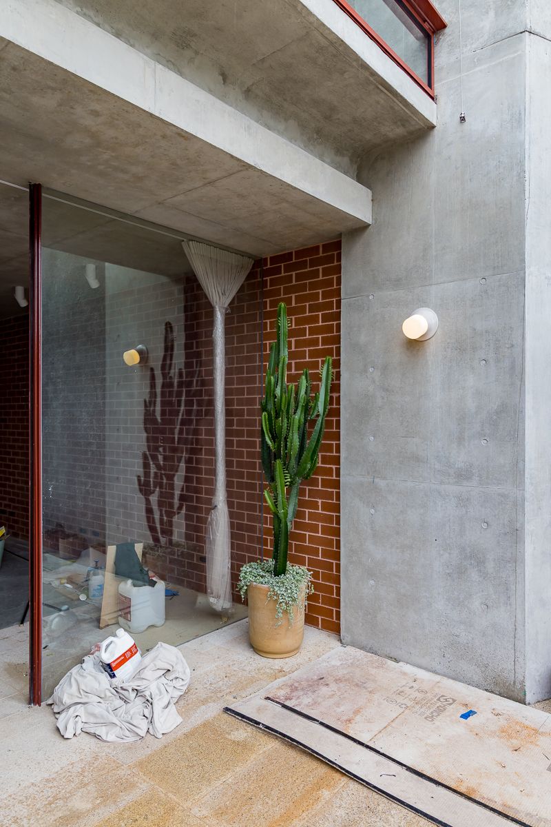 Entrance corner of a concrete and brick home with large glass doors, a tall cactus in a pot and protective floor coverings.