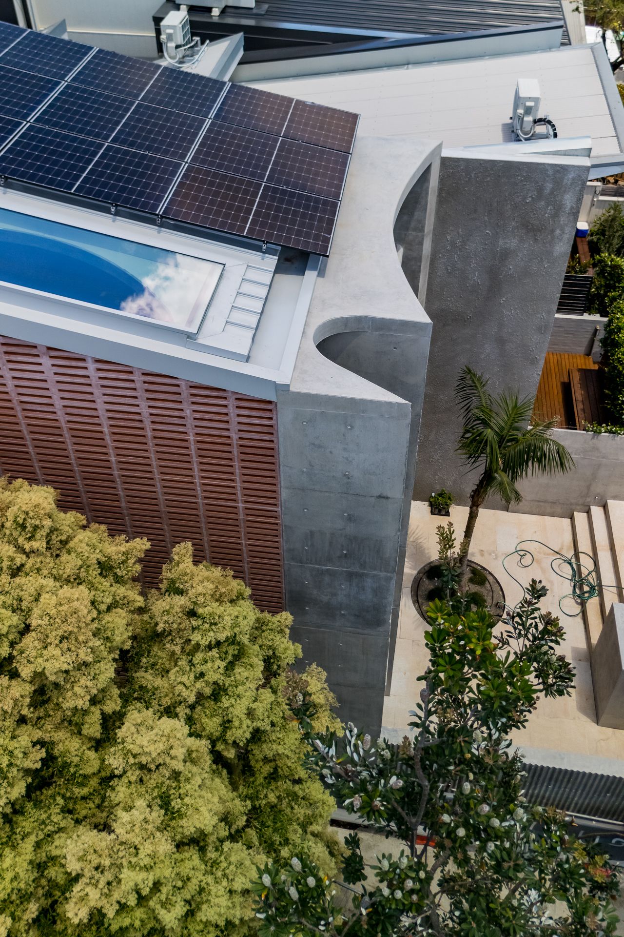 Aerial view of a modern concrete house with rooftop solar panels, a skylit pool, a timber-screened facade and lush green trees.