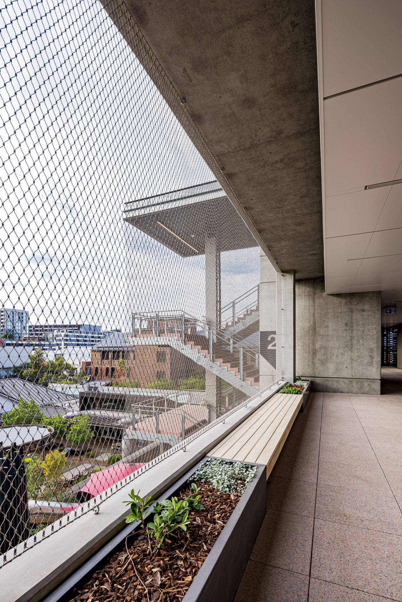 Open-air concrete balcony with a wooden bench, planter of greenery, mesh safety netting and an exterior staircase overlooking city rooftops.