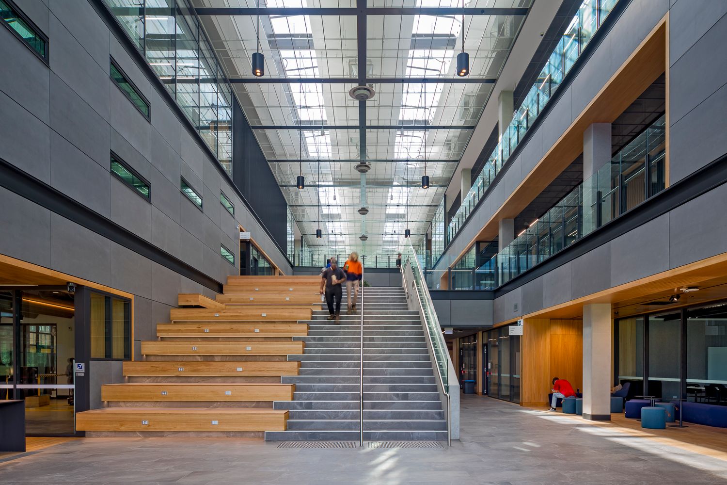 Students walk toward a wide staircase with timber seating steps in a bright multi-level atrium with a skylit ceiling and glass balconies.