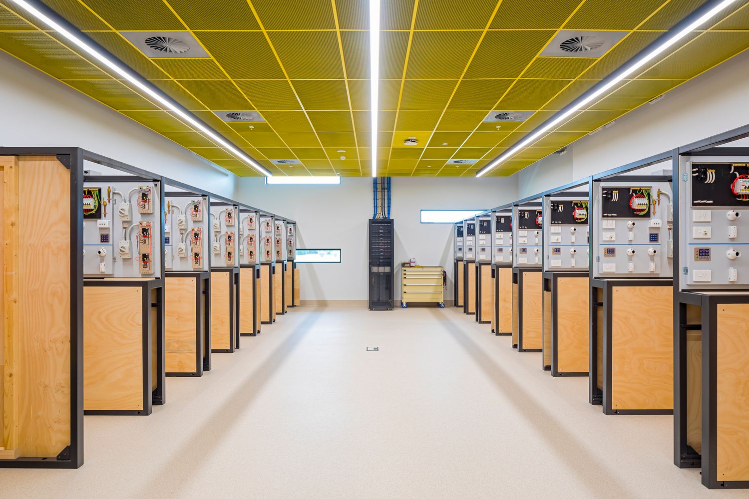 Electrical training lab with rows of timber-backed test panels and a central aisle beneath yellow ceiling panels.