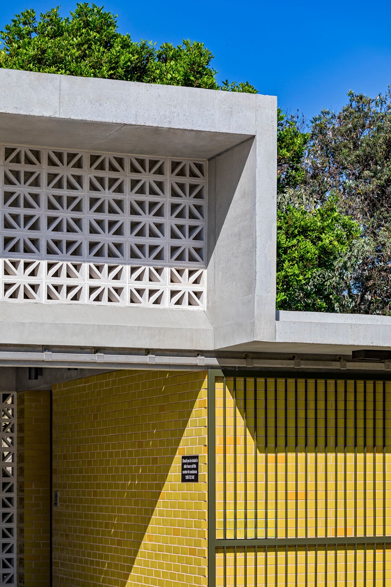Modern concrete building with a geometric breeze block screen above a yellow tiled brick wall and slatted gate, framed by green trees.