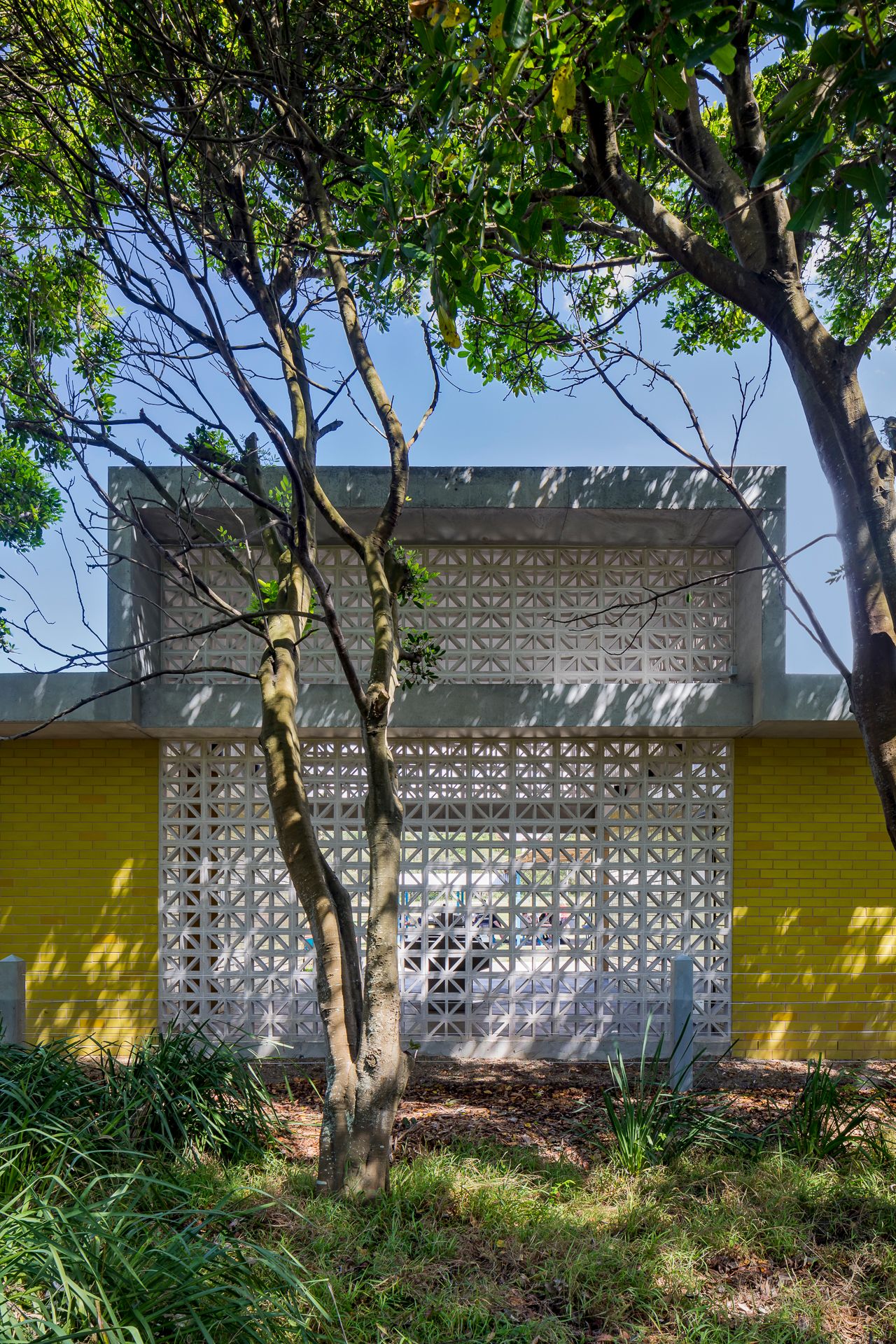 Exterior of a restroom building with a white patterned breeze block screen and yellow brick walls framed by slender tree trunks and foliage.