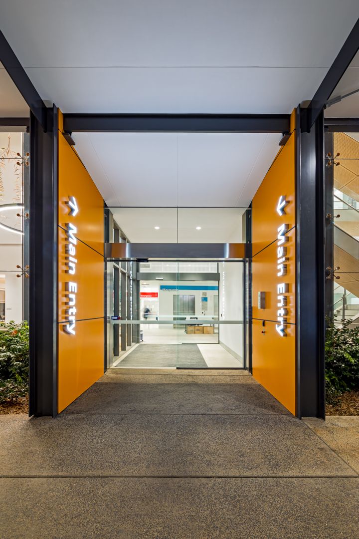 Bright orange hospital entrance with illuminated Main Entry signs and glass doors leading to a modern, well-lit interior lobby.
