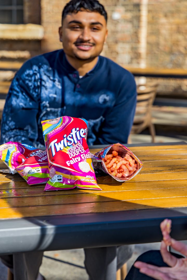 Smiling young man at an outdoor wooden table with packets of Twisties Fairy Floss crisps and spilled cheese snacks.