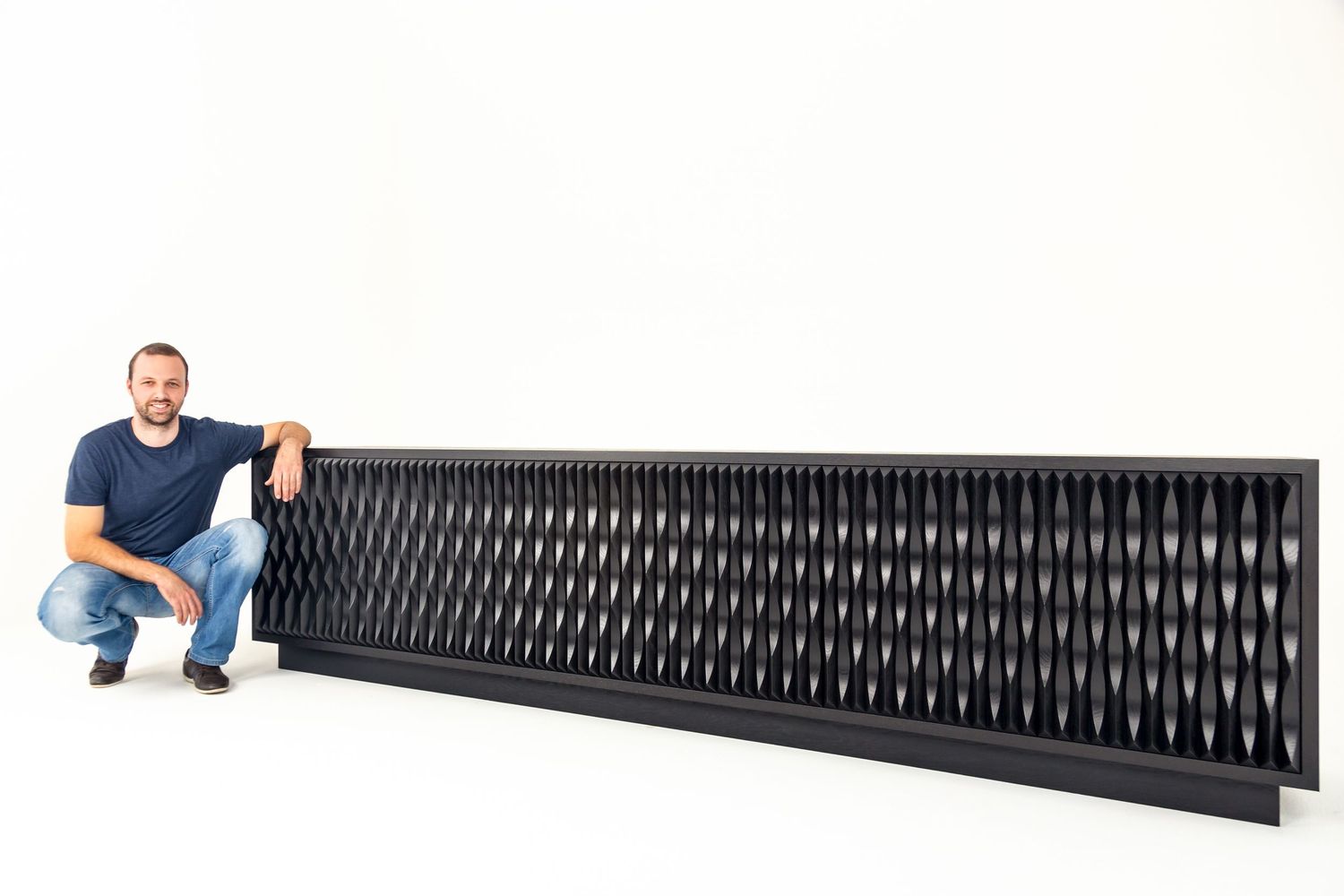 Smiling man in a navy t-shirt crouches beside a long black sculptural panel with a textured wave pattern against white.