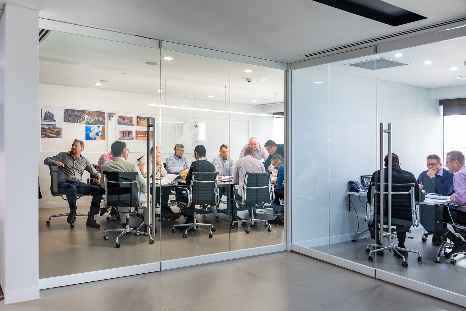 Group of professionals meets around a conference table in a glass-walled modern office meeting room with a person presenting.