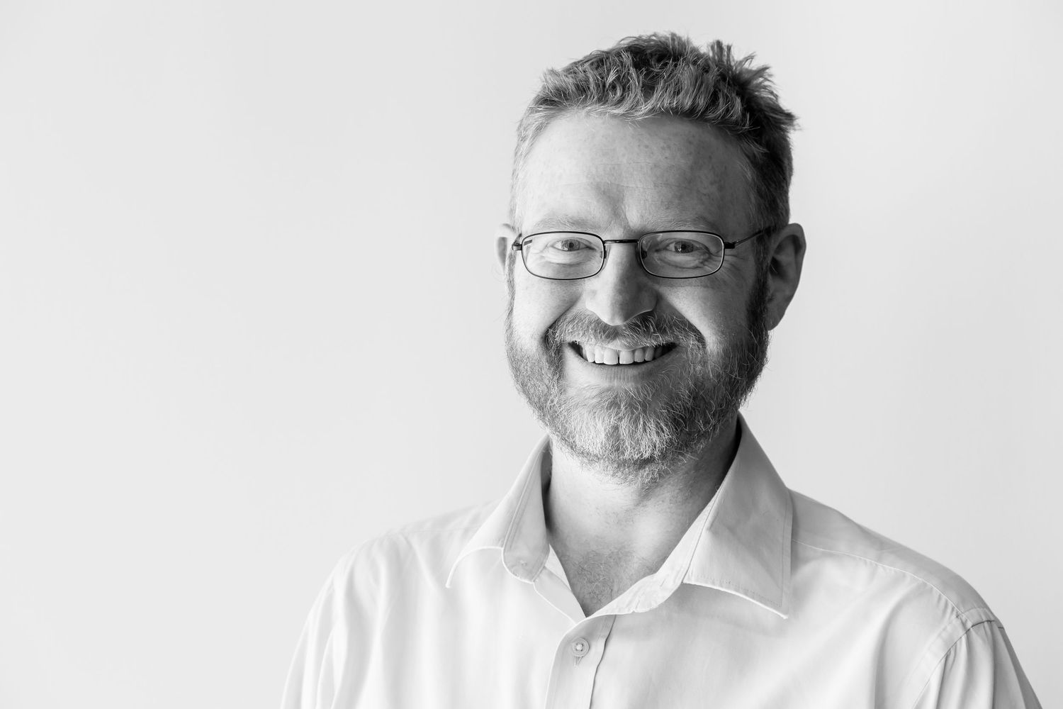 A black and white portrait of a smiling bearded man wearing glasses and a white collared shirt against a plain light background.