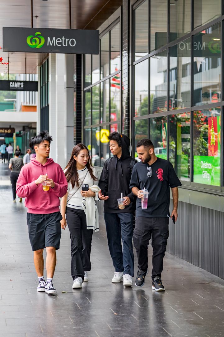 Four young friends walk together carrying drinks along a covered shopping walkway beneath a Metro supermarket sign.