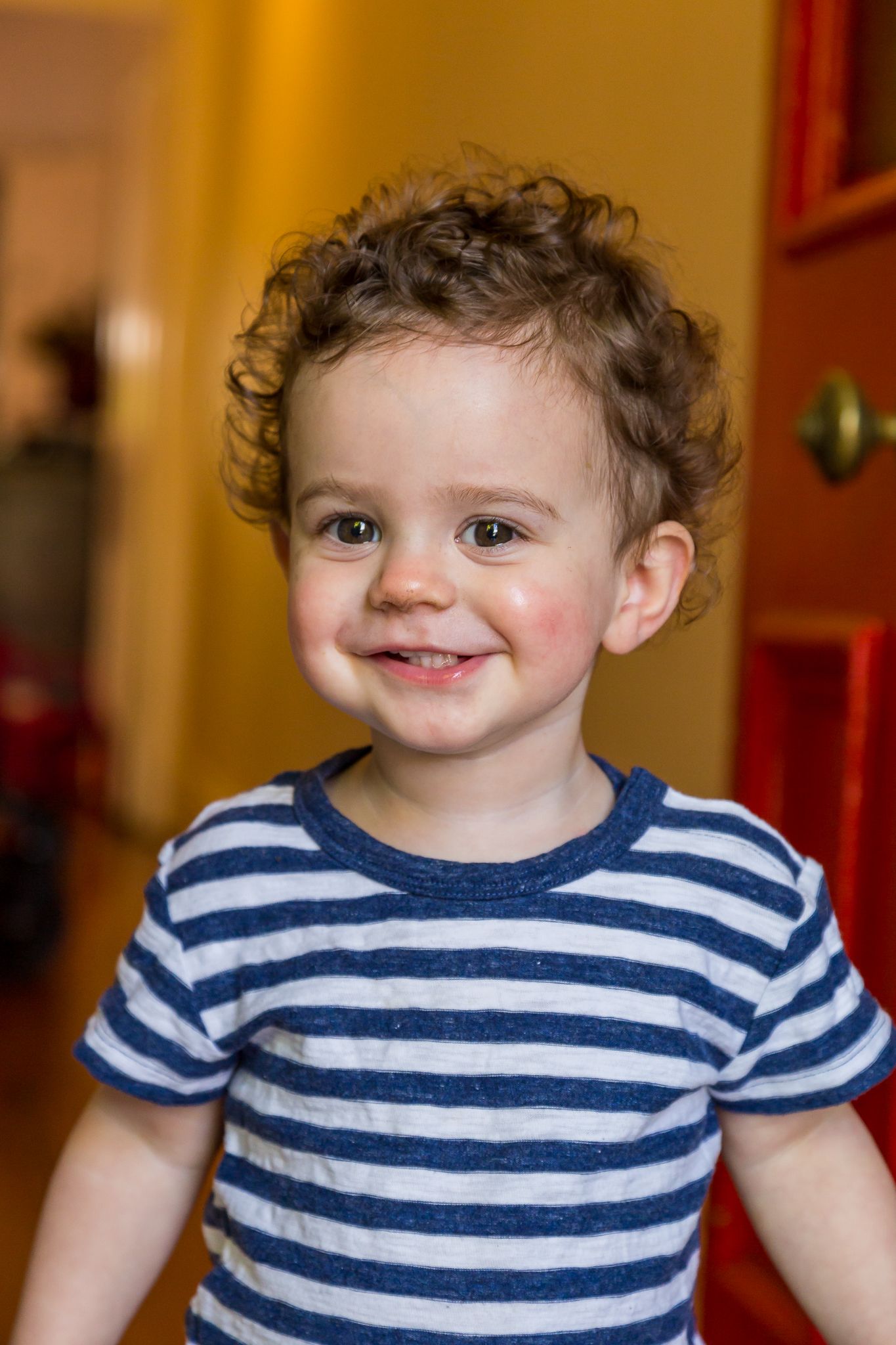 Smiling toddler with curly brown hair and rosy cheeks wears a navy and white striped t-shirt in a warm indoor hallway.