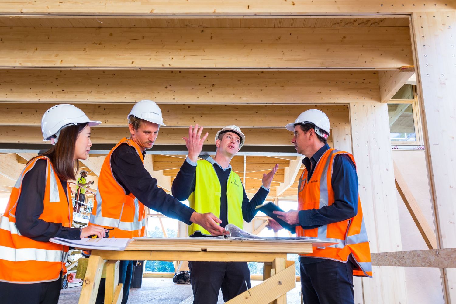 Four construction professionals in hard hats and high-visibility vests discuss building plans at a timber-framed site.