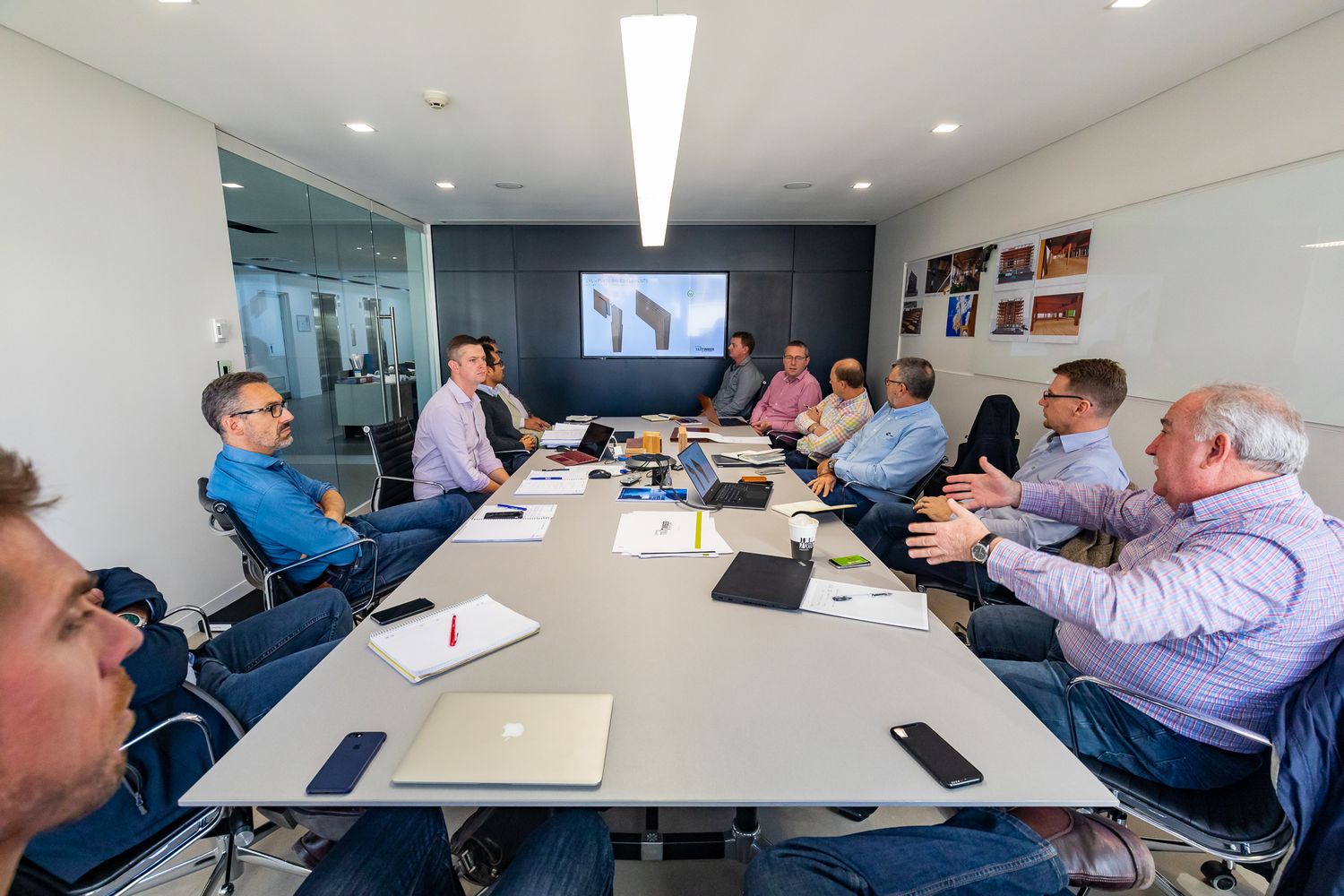 A group of men sit around a glass meeting table with laptops and notebooks in a bright modern conference room.