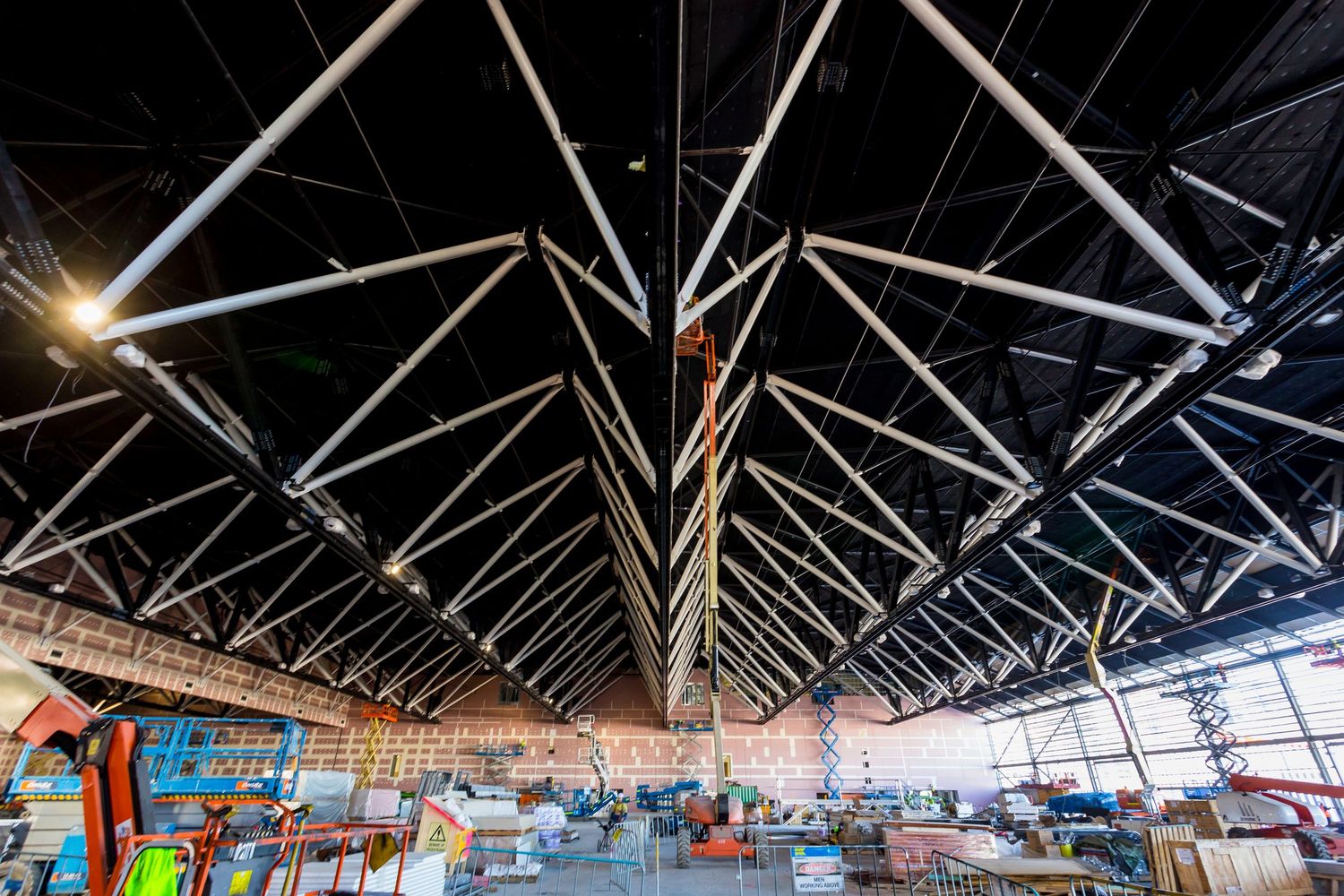 Interior of a large arena under construction with exposed steel roof trusses, scaffolding, and scissor lifts working across the open floor.