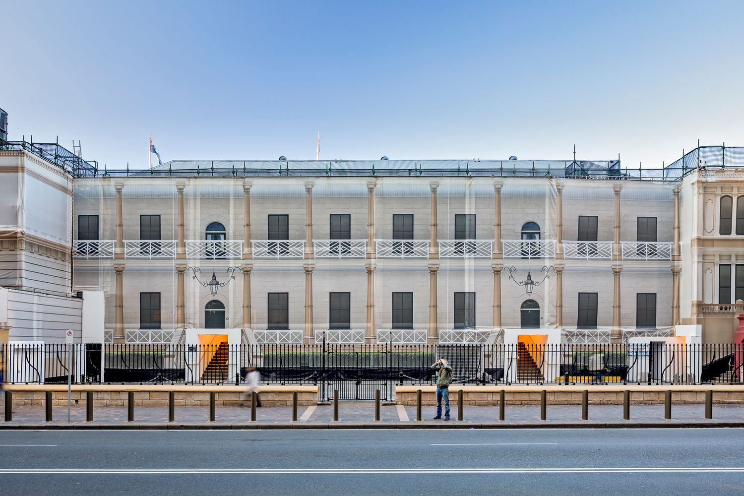 Wide view of a long historic building facade under restoration with scaffolding, balconies and a pedestrian standing on the footpath.