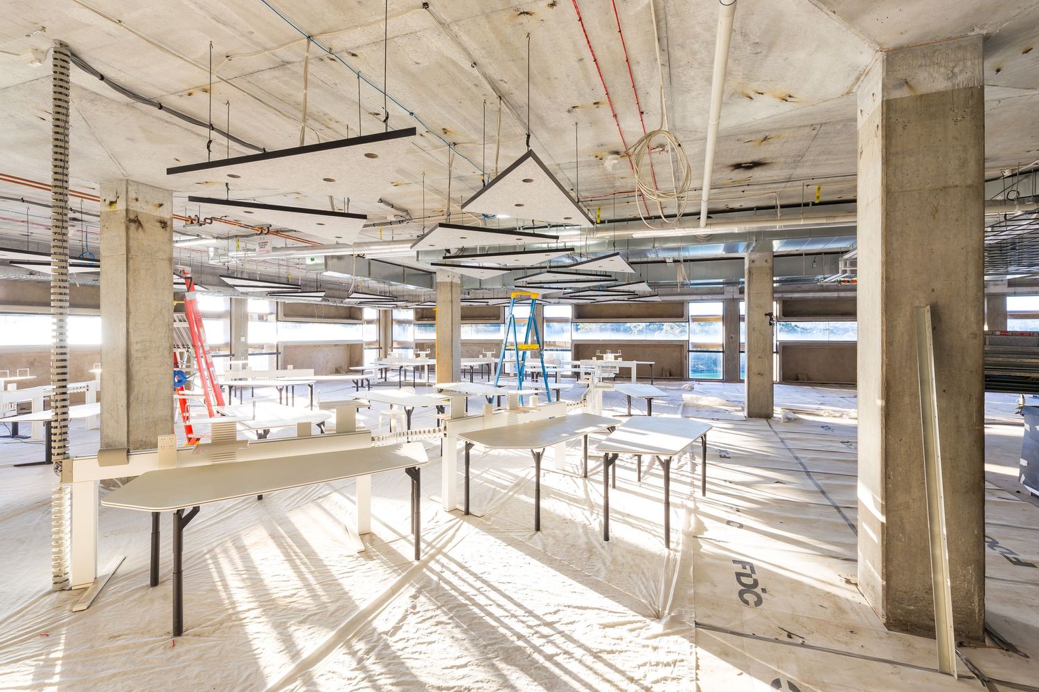 Spacious unfinished interior floor with concrete columns, exposed ceiling services and temporary work tables lit by natural light.