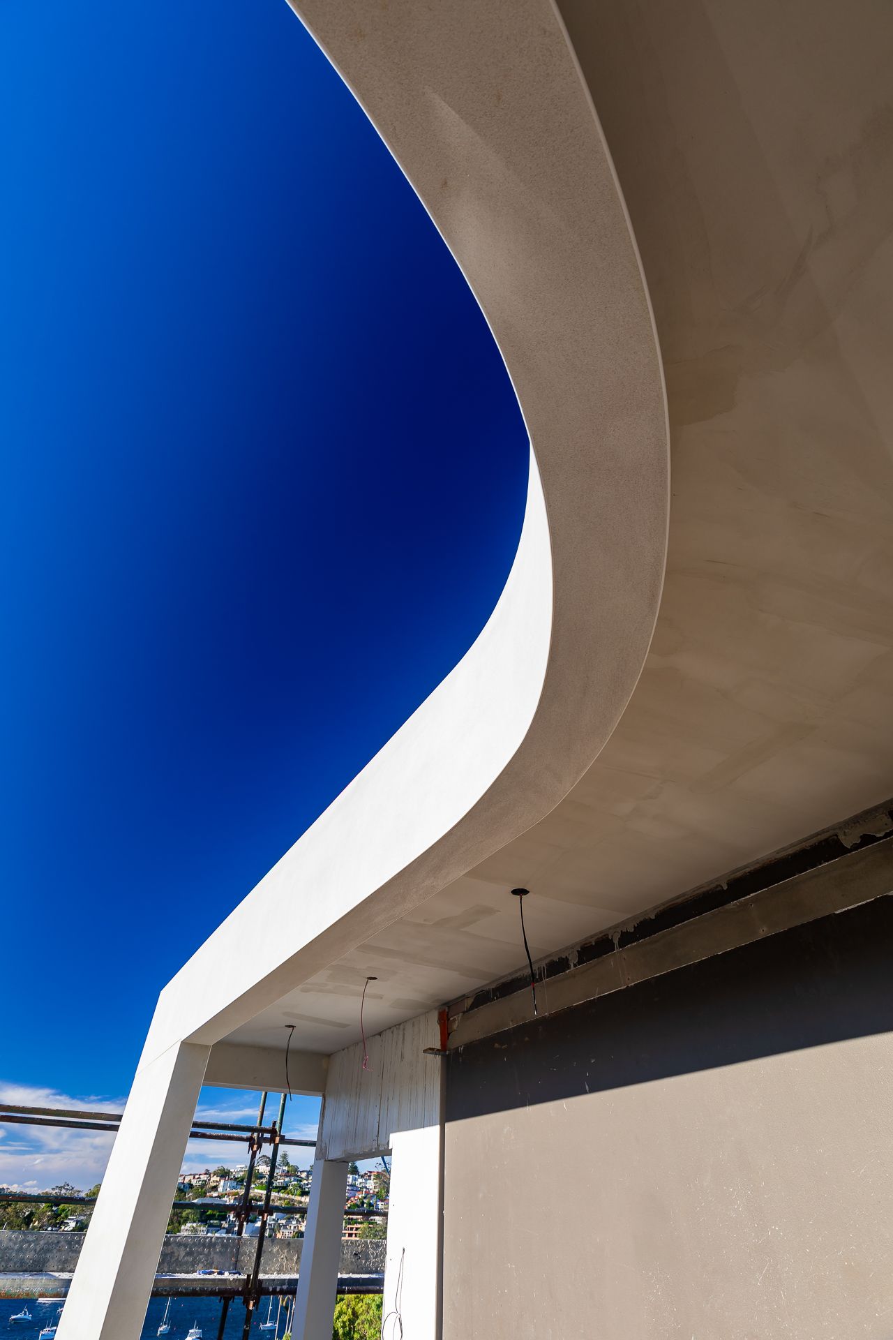 Sweeping curved white concrete soffit and column against a vivid blue sky, with harbour water and scaffolding visible below.