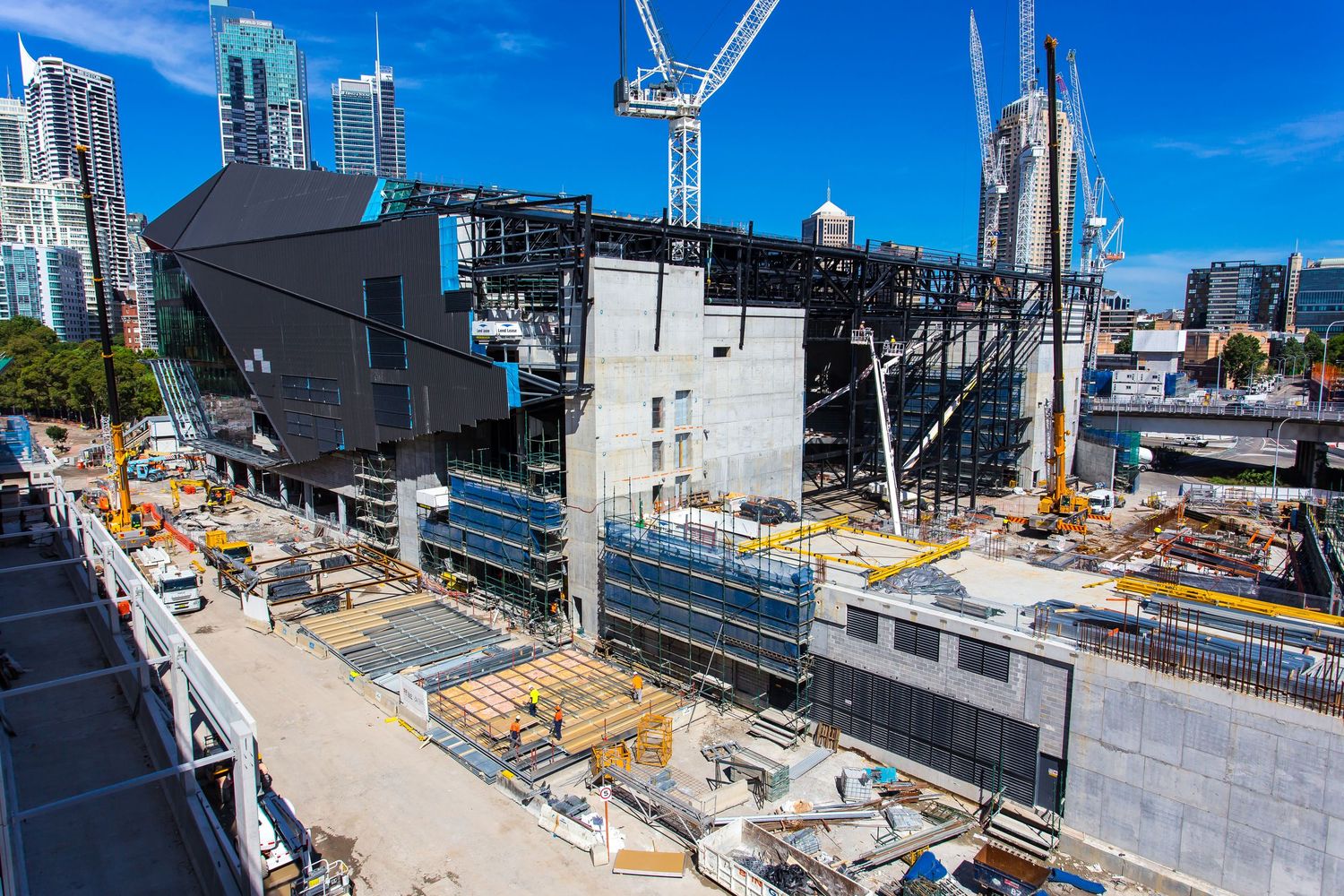 Large construction site in a city with cranes, steel framing, scaffolding and partially built concrete structures beside a river.