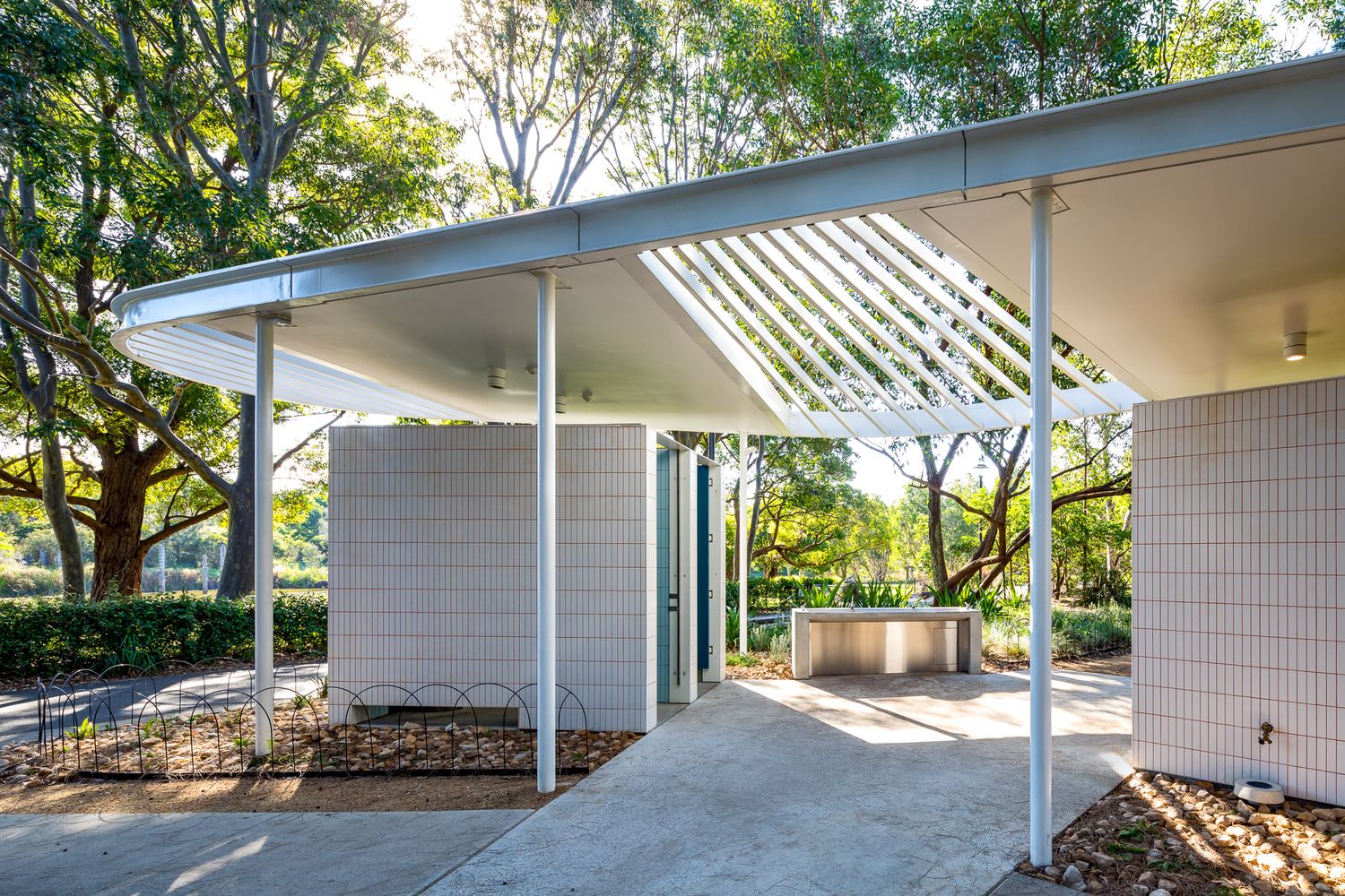 Modern white pavilion with a slatted louvred roof and brick walls beside a paved path, surrounded by tall trees.