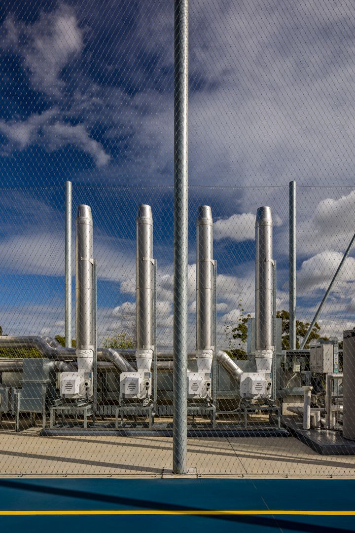 Row of silver industrial exhaust flues and ductwork behind a wire mesh fence against a blue sky with scattered clouds.