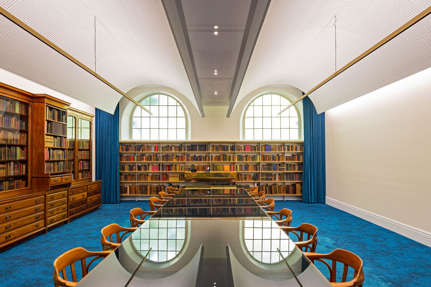Grand library reading room with a long polished table, wooden chairs, arched windows, built-in bookshelves and bright blue carpet.
