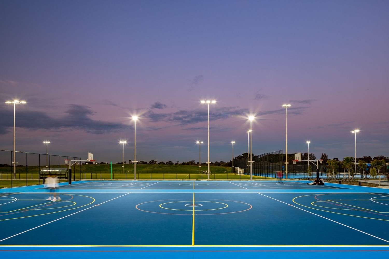 Blue outdoor netball courts lit by tall floodlights at dusk under a purple sky, with green playing fields in the distance.