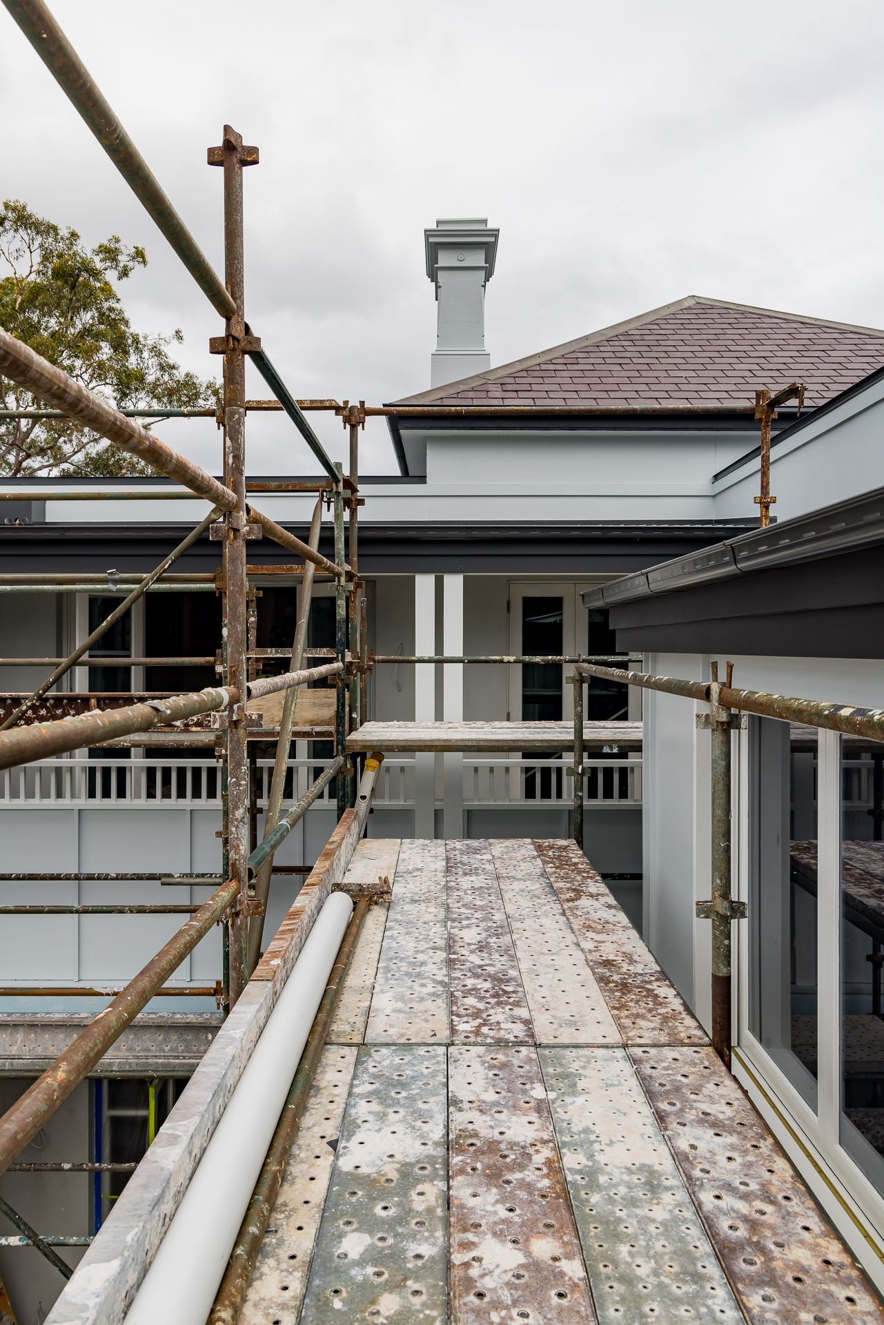 Scaffolding with perforated planks beside a grey-painted heritage home with a slate roof and chimney during exterior renovation.