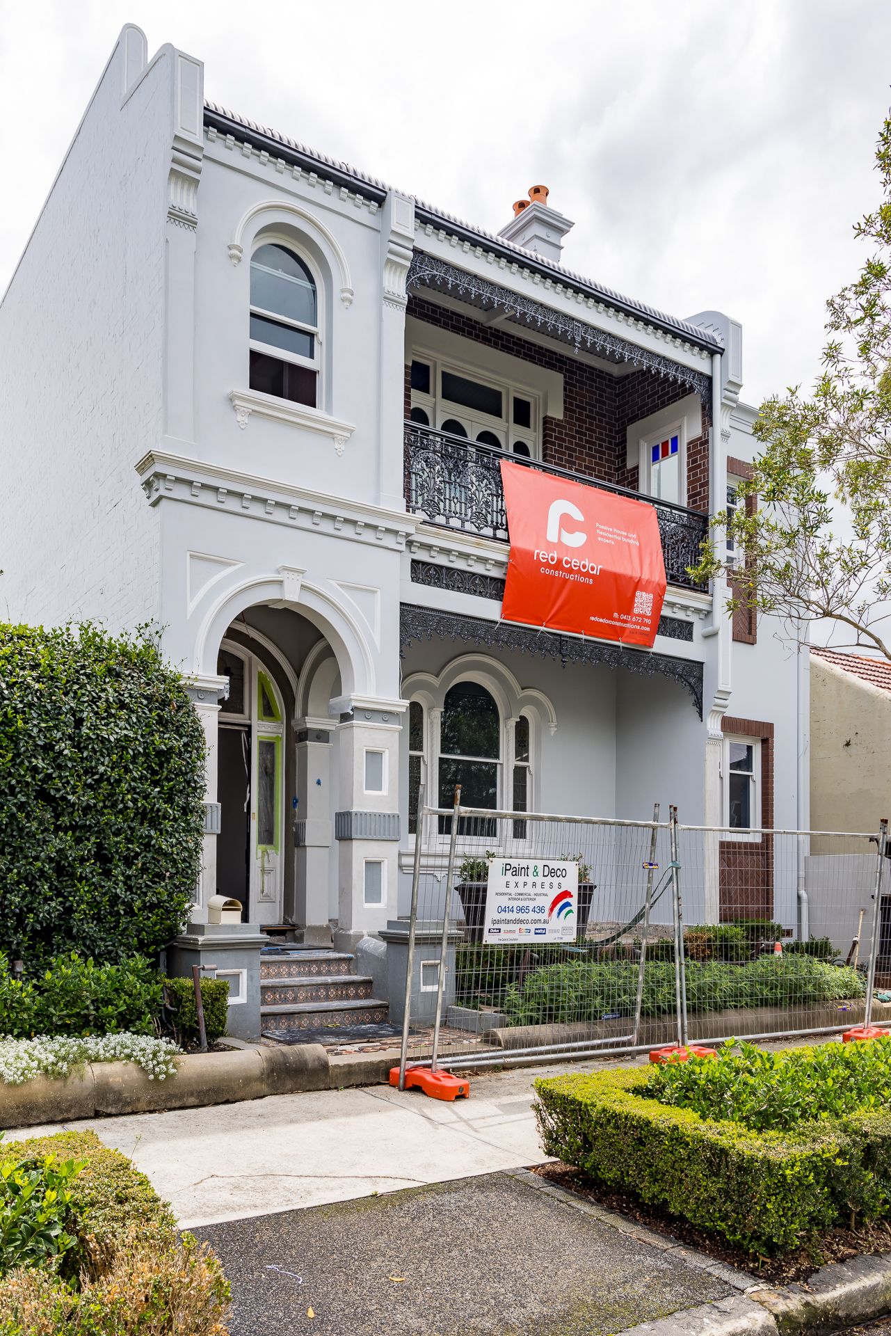 Two-storey Victorian terrace with a cast iron lace balcony, arched entry and a Red Cedar Constructions banner during renovation.