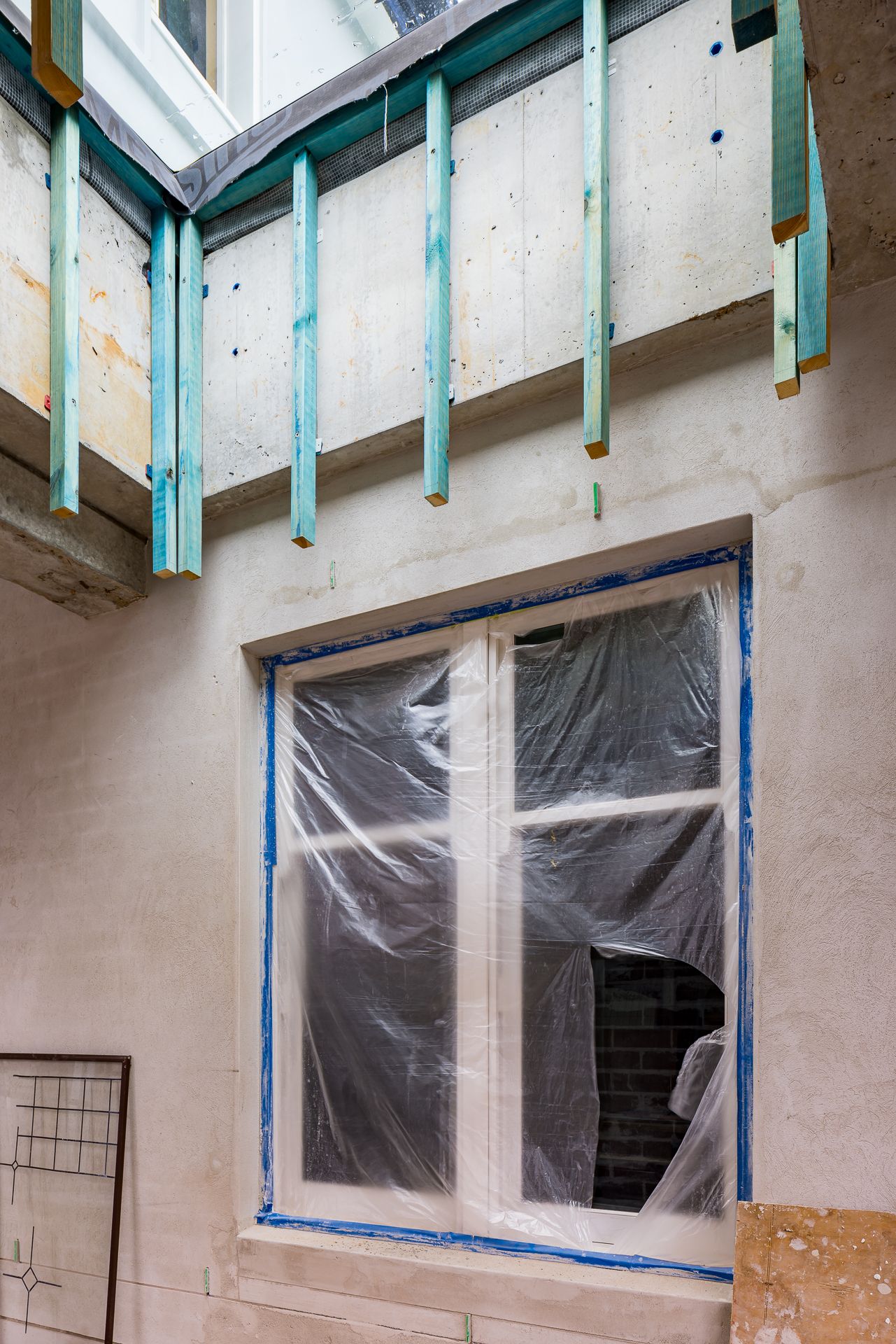 Exterior wall under construction with a window protected by plastic sheeting and blue tape, plus teal-painted timber framing above.
