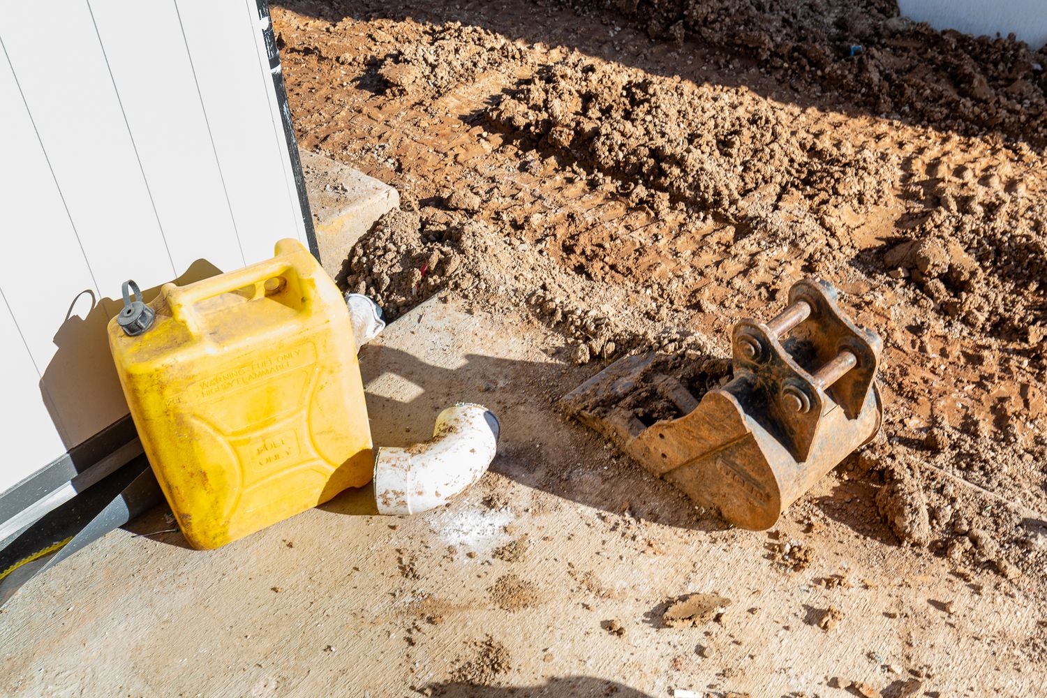 Yellow plastic jerry can, excavator bucket, and a white pipe fitting on churned dirt beside a building wall on a worksite.