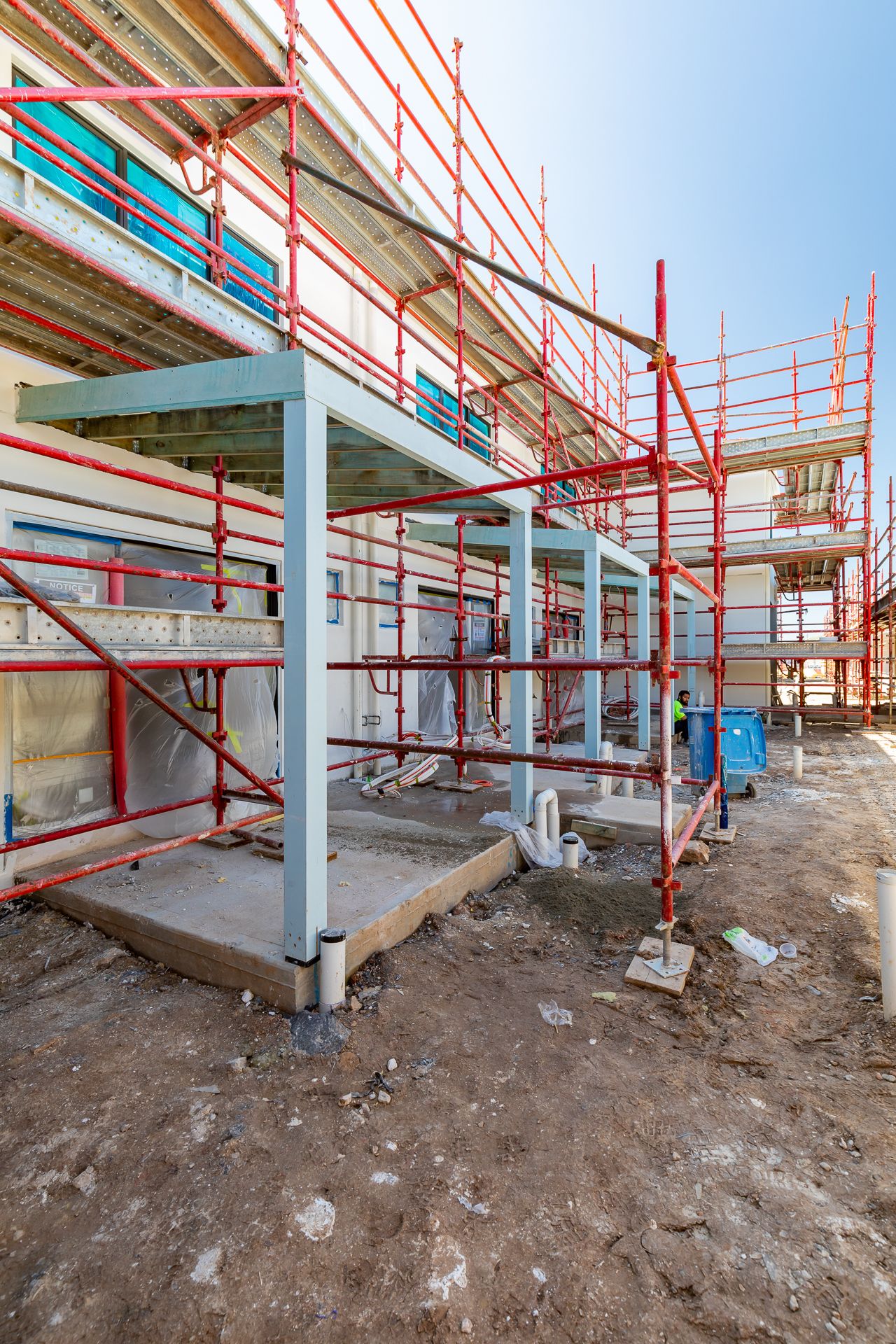 Red scaffolding surrounding a multi-storey building under construction with steel support columns and a concrete slab on a dirt site.