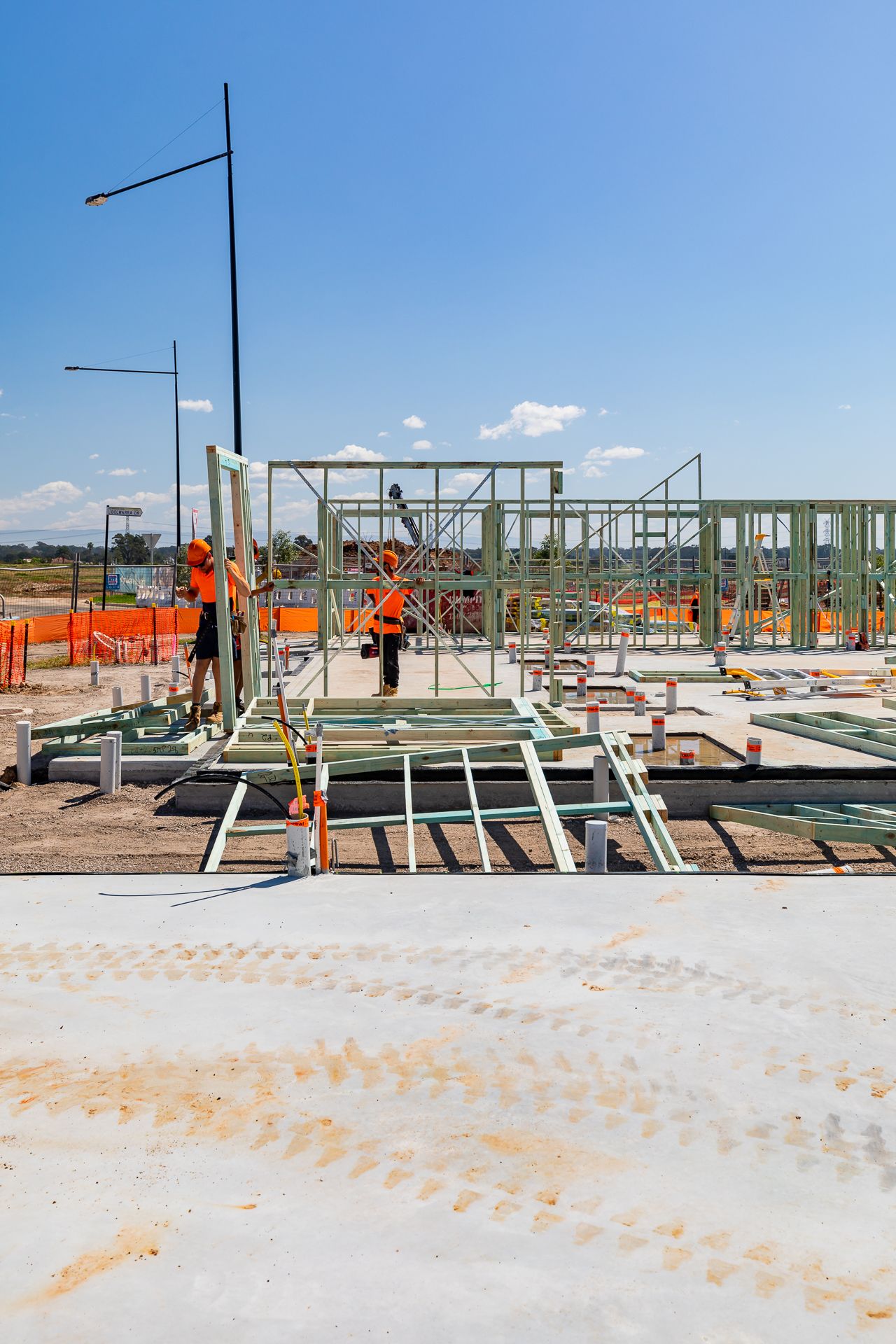Construction workers in orange high-vis clothing assembling steel wall frames on a concrete slab at a new housing development.