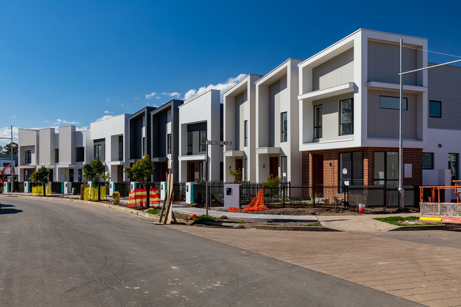 Row of modern white and grey townhouses with balconies on a newly built street under a clear blue sky.