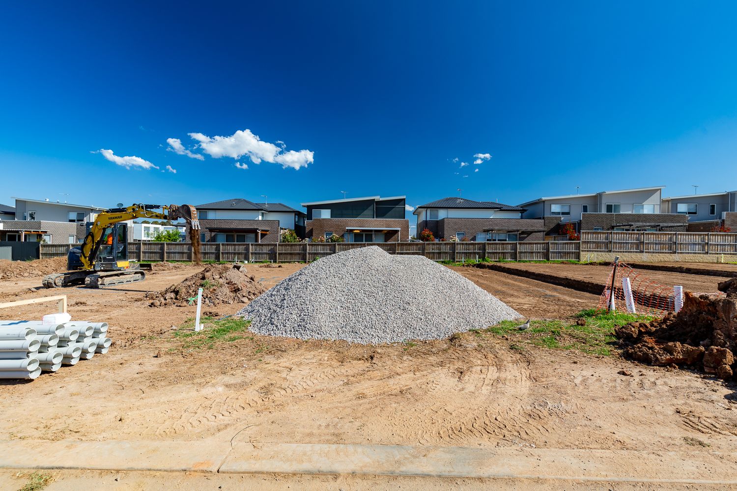 Excavator and gravel pile on a graded building site with new suburban houses and a fence in the background.