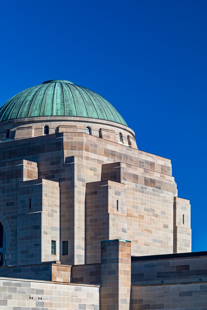 Green copper dome atop a sandstone memorial building with stepped stone walls against a deep blue sky.