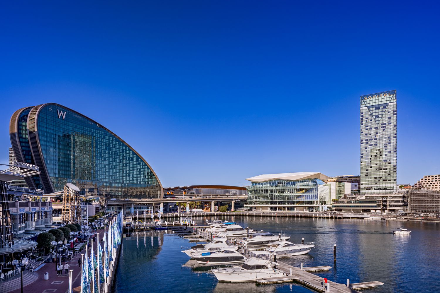 Modern glass-domed waterfront building and high-rise tower beside a marina with moored yachts under a clear blue sky.