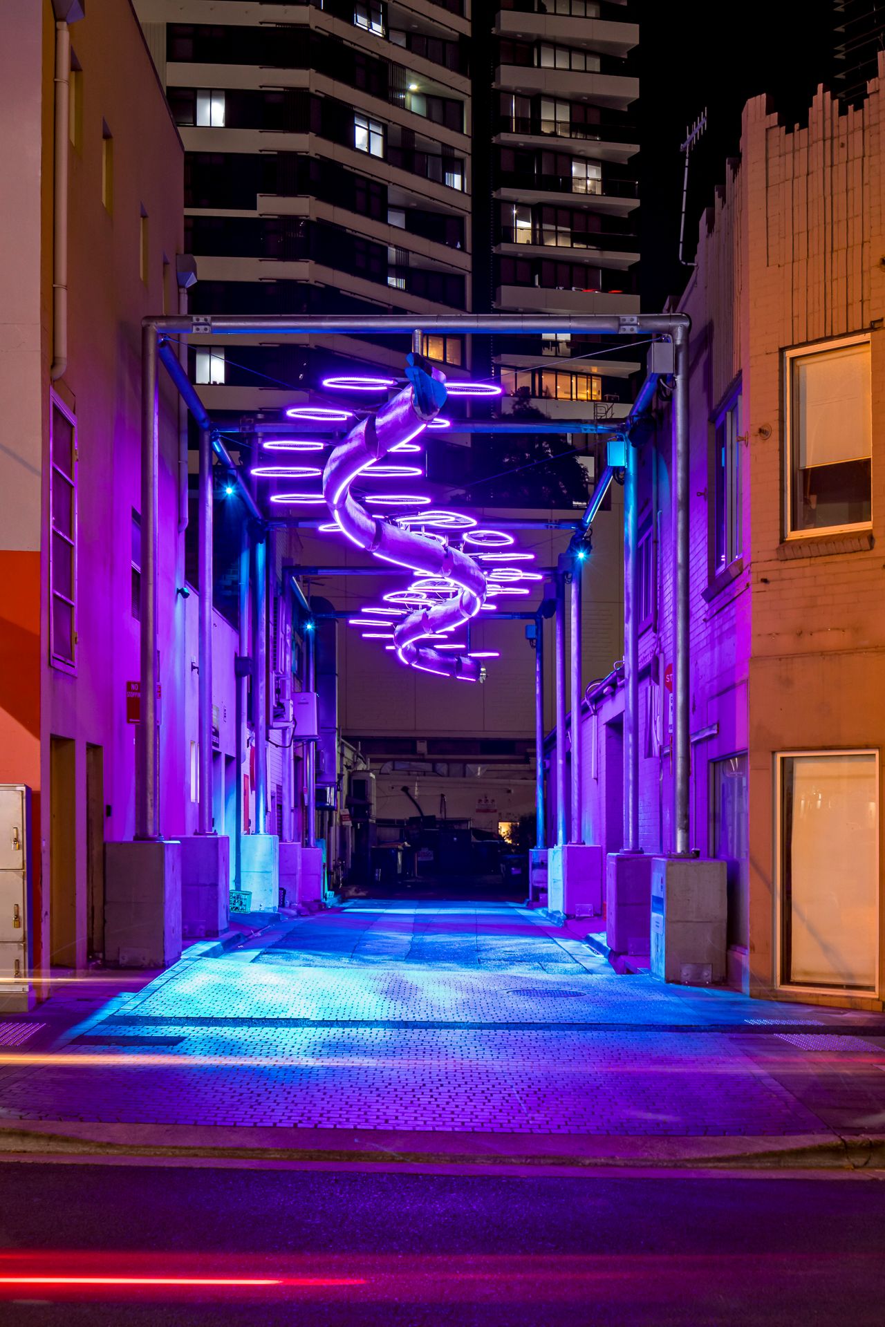 Vivid purple neon spiral sculpture suspended over a empty city laneway at night, reflecting on the blue-lit pavement below.