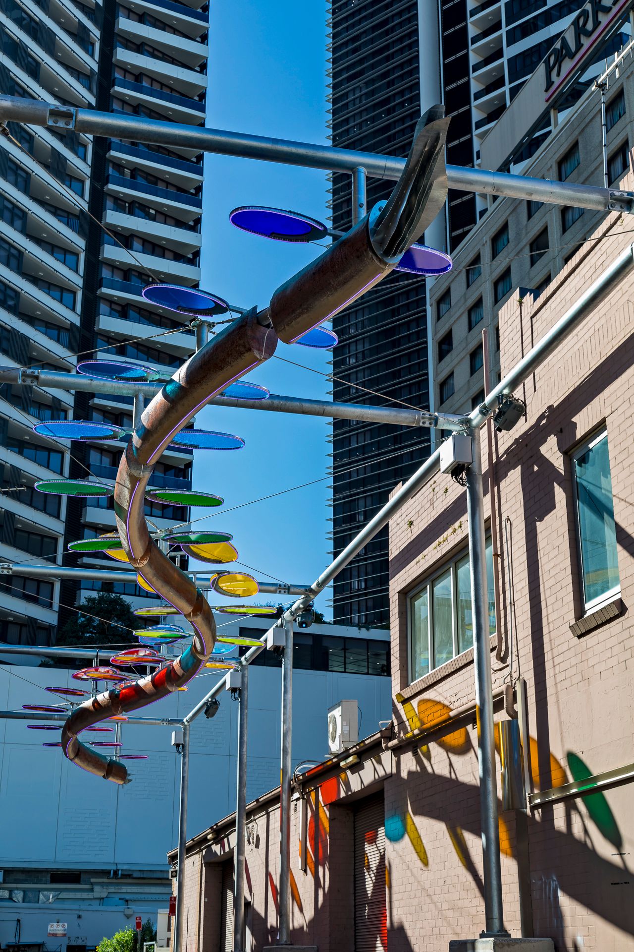 Winding sculpture with blue, green and orange discs above a brick laneway building, with skyscrapers in the background.