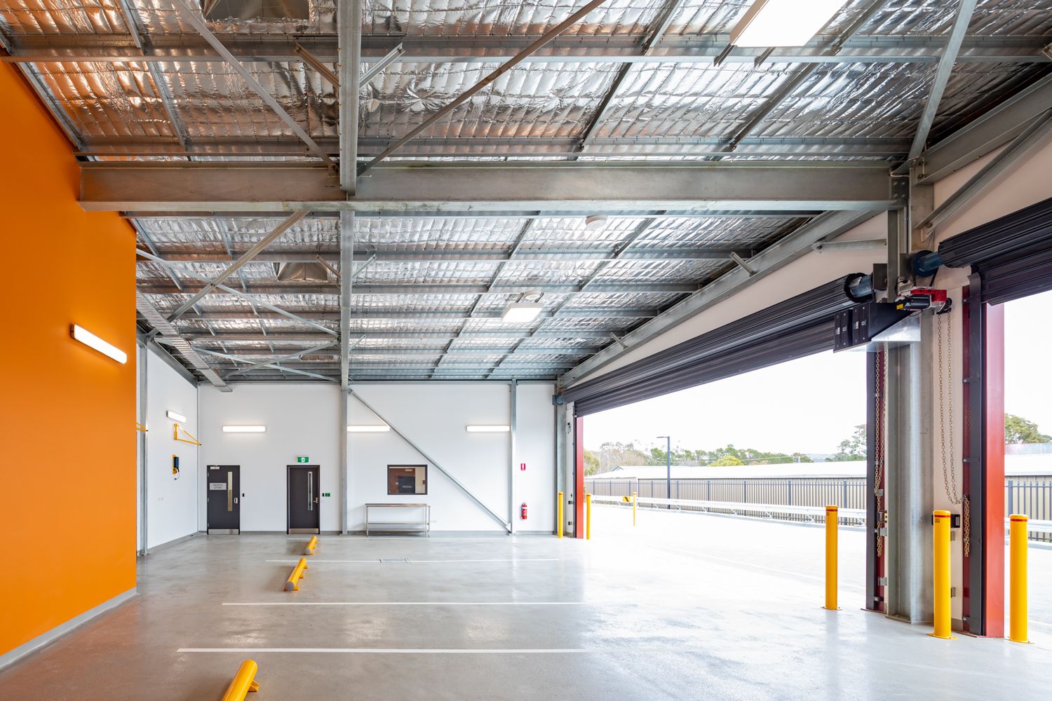 Spacious industrial warehouse interior with exposed steel roof trusses, orange feature wall and yellow safety bollards.