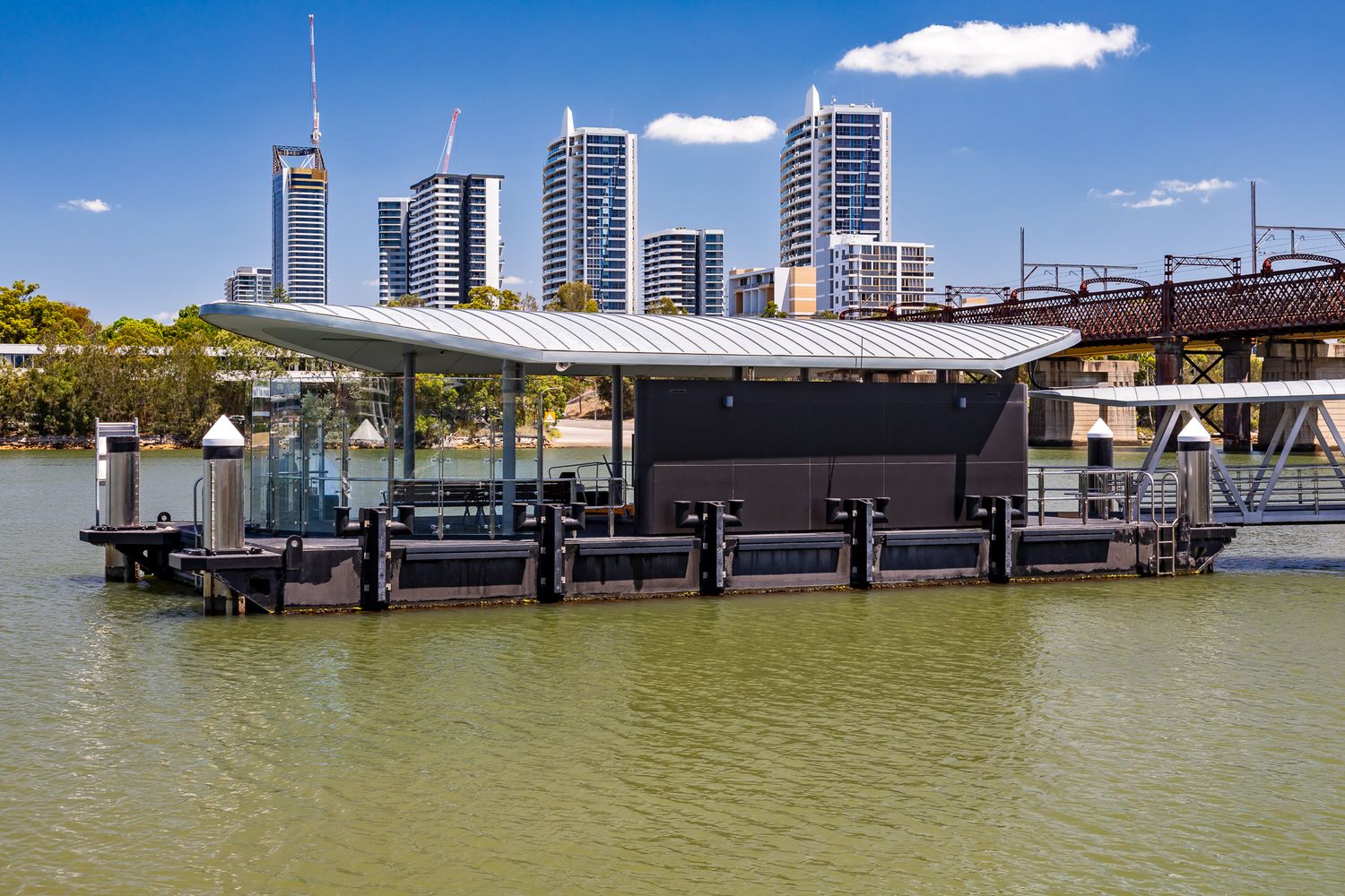 Dark-clad ferry pontoon with a flat canopy on a green river, with a steel bridge and tall city apartment towers in the background.