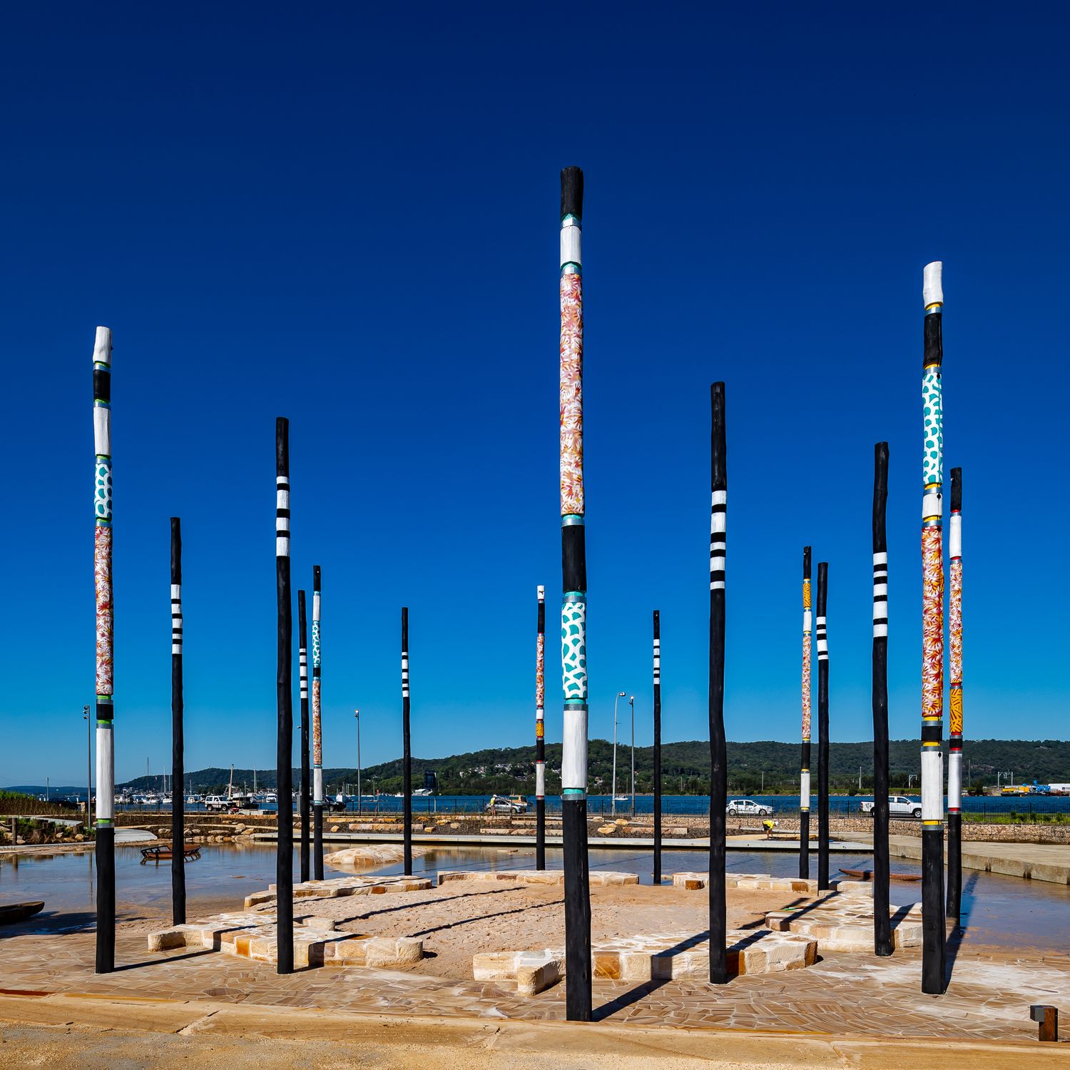 A cluster of tall painted poles with patterned bands stands over a sandstone water feature near the waterfront.