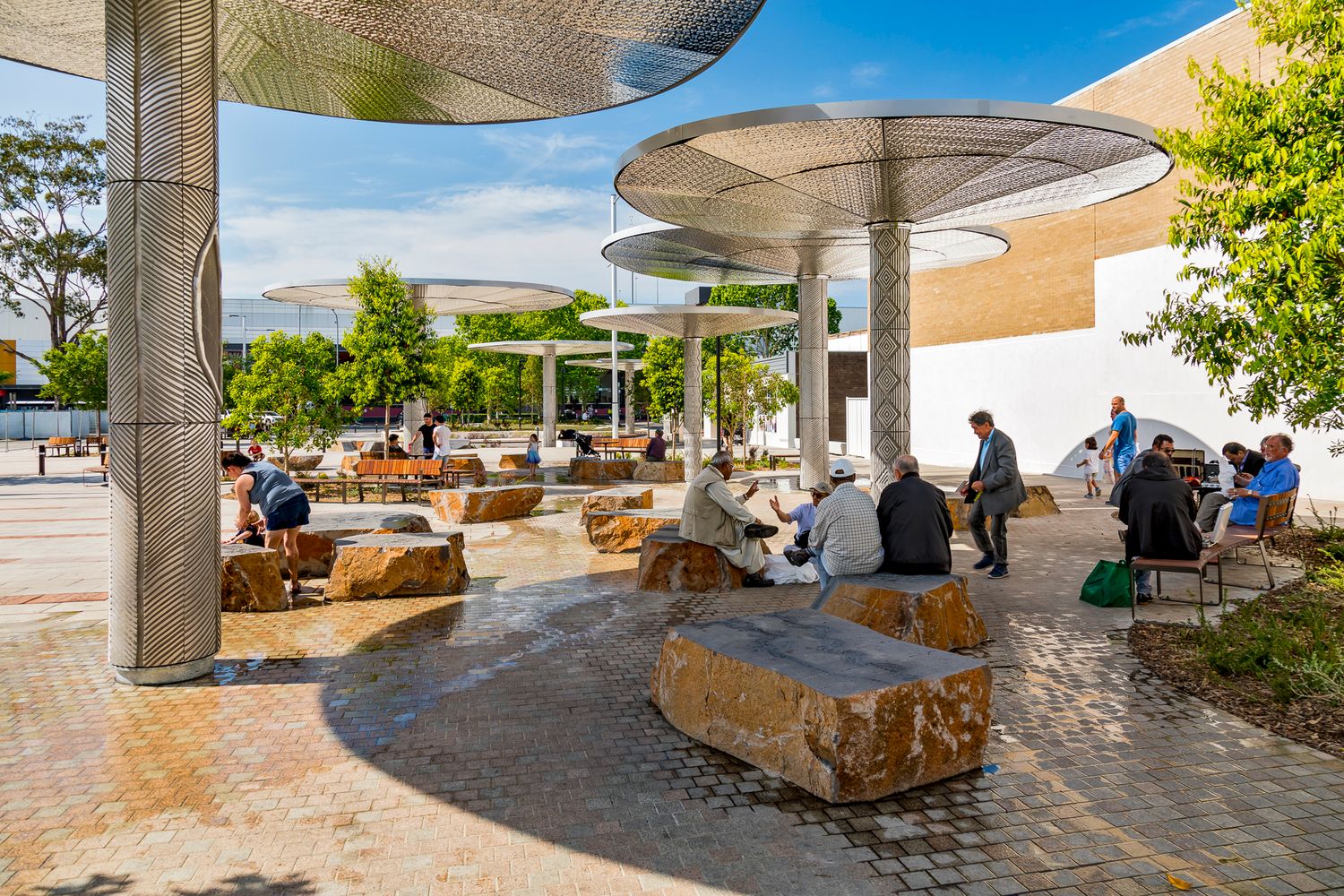 People gather on large sandstone boulder seats under modern metal canopies in a sunlit public square with trees and paving.