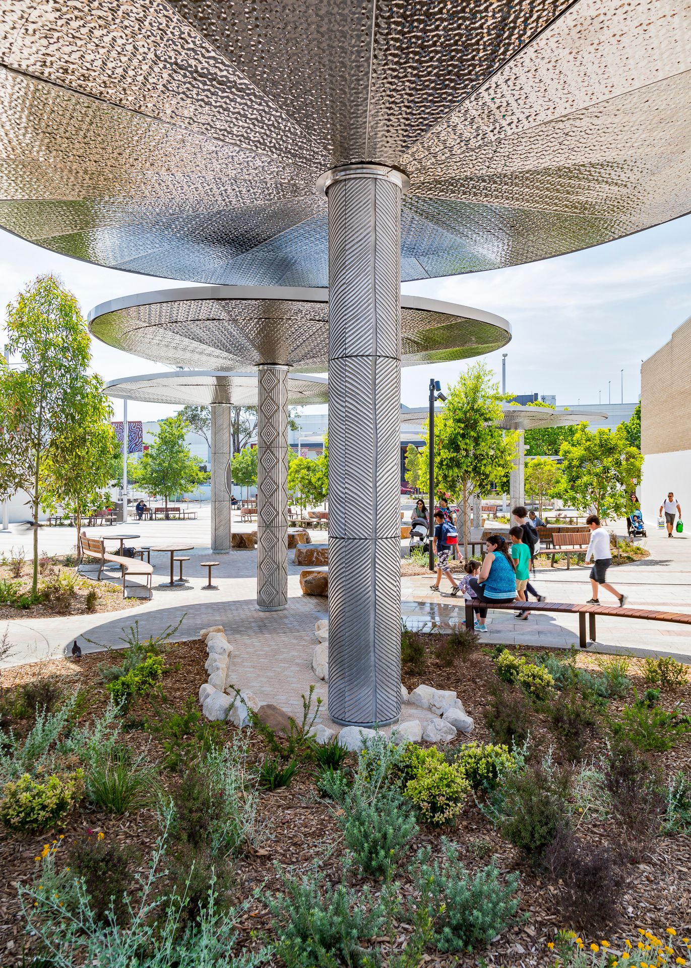 Patterned metal column and perforated canopy rise above native garden plants as visitors sit on benches in a public plaza.