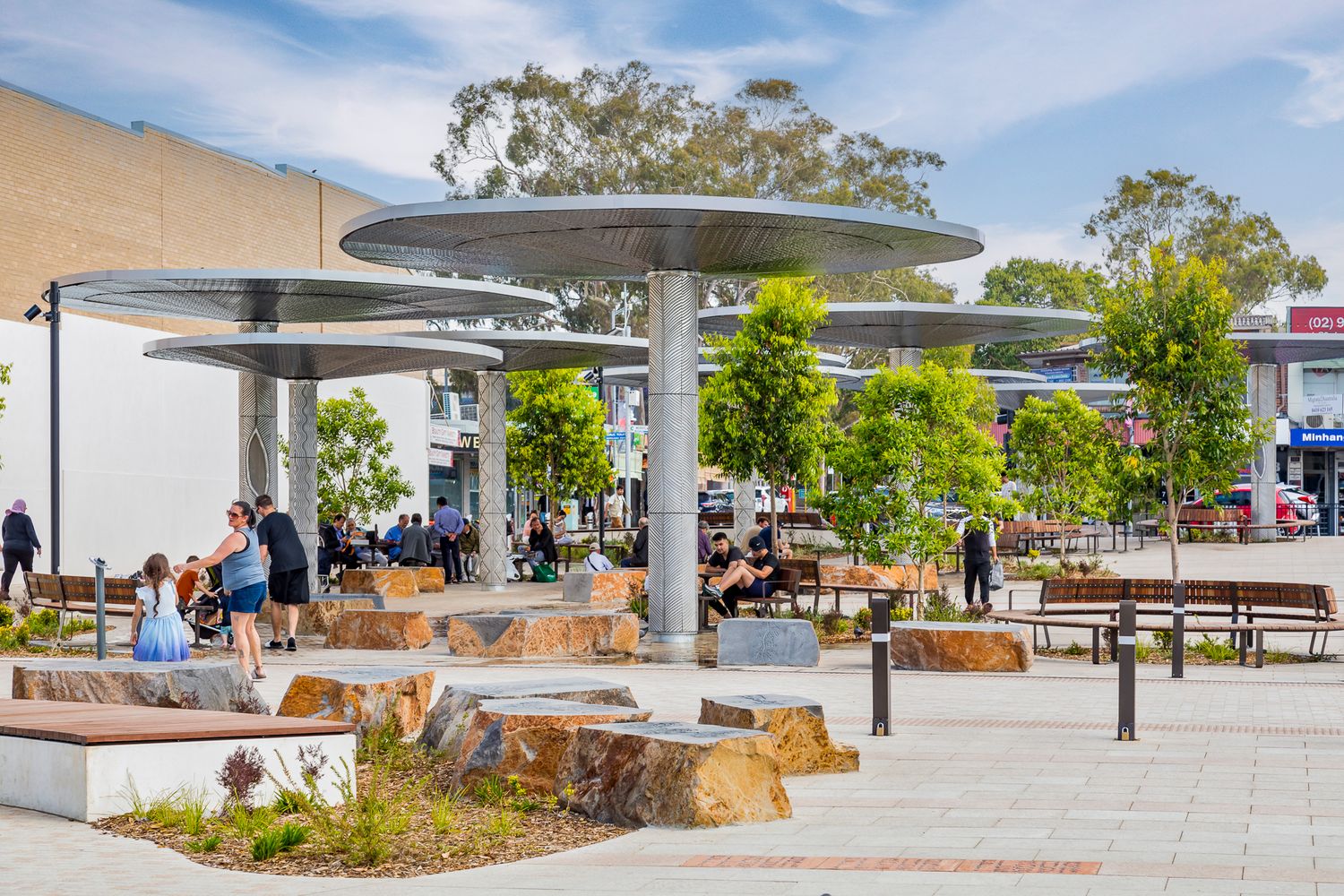 Visitors relax on benches and boulder seating beneath large mushroom-shaped metal canopies in a sunny public plaza lined with young trees.