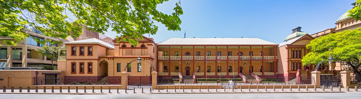 Panoramic view of a historic sandstone building with a double-storey verandah, red brick wings and tree-lined street frontage.