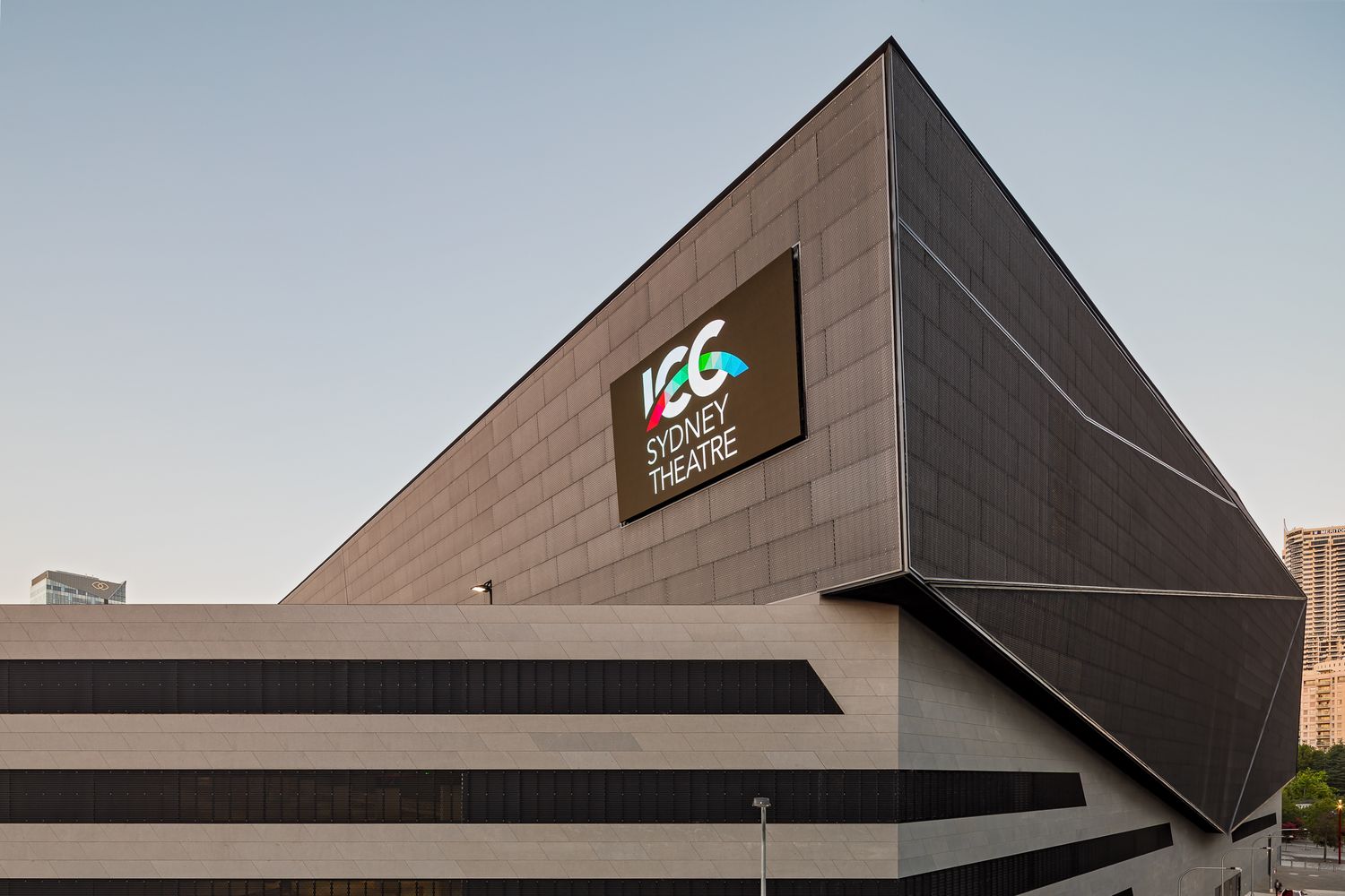 Angular dark facade of the Sydney Theatre building with an illuminated sign against a soft evening sky.