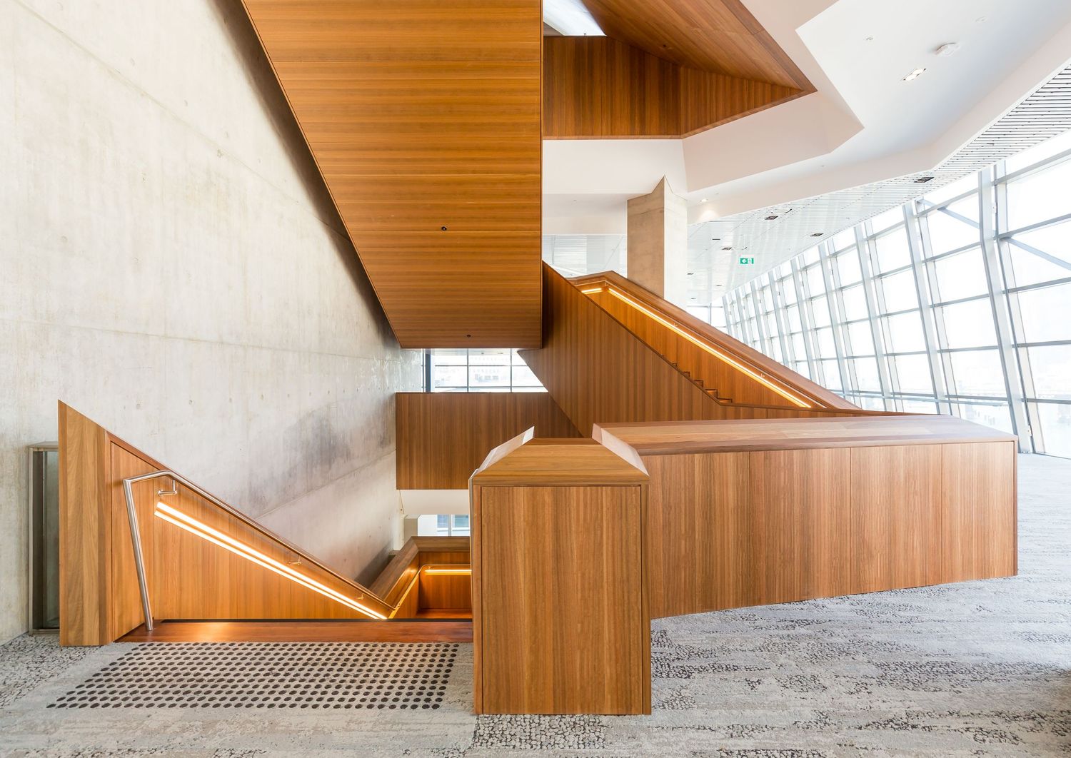 Timber-clad staircase with integrated lighting beside concrete walls and large windows in a modern interior.